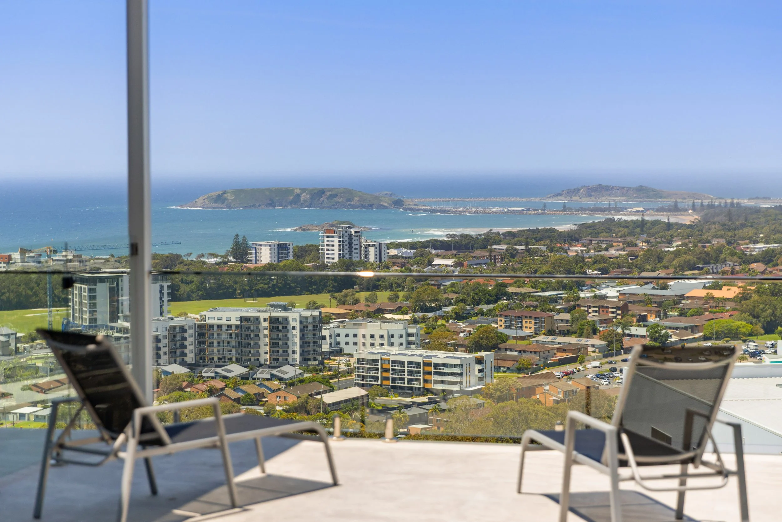 Balcony with two chairs overlooking a city and coastal landscape with buildings, greenery, and the ocean in the background.