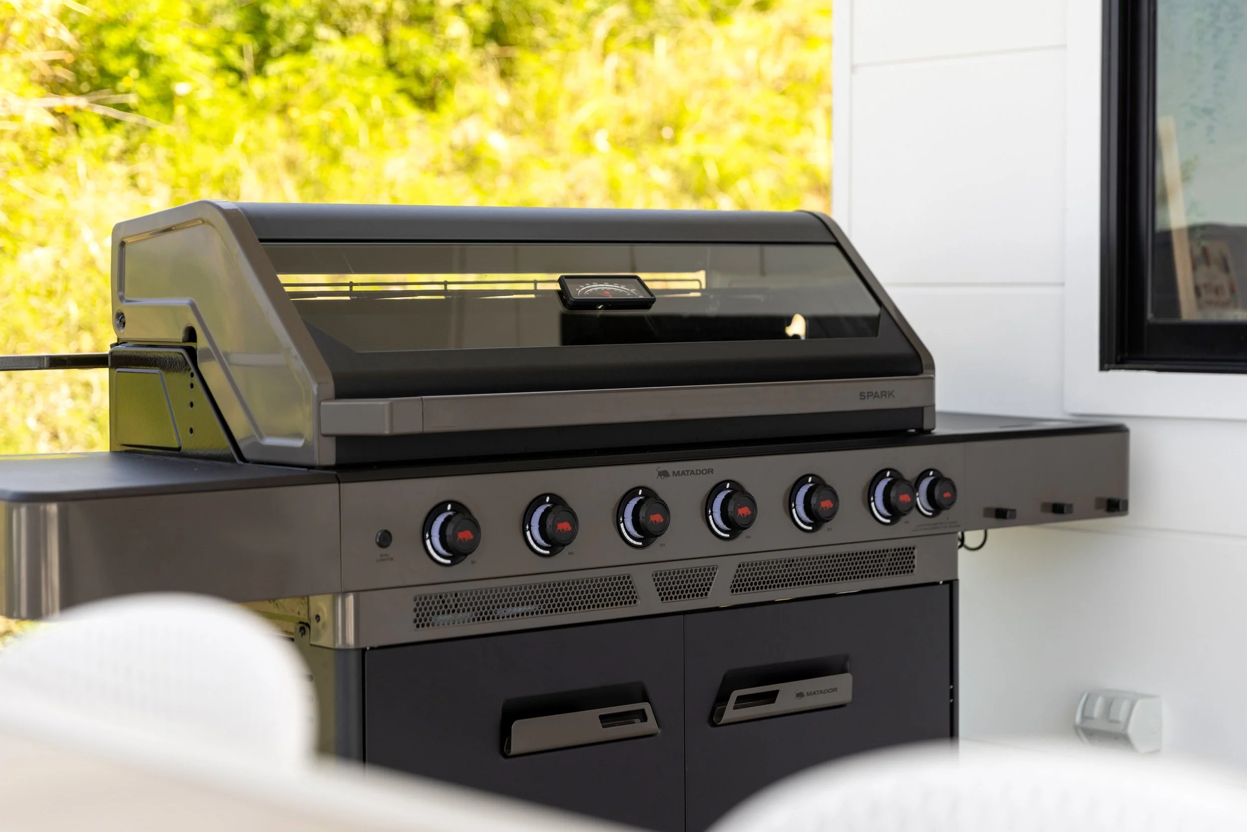A modern outdoor grill with a glass lid, control knobs, and a built-in thermometer, positioned against a white wall with a window showing greenery outside.