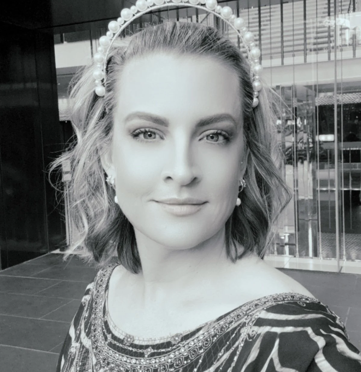 A woman with shoulder-length wavy hair, wearing a pearl headband and earrings, taking a selfie outside a modern building with glass windows. She has light-colored eyes and is dressed in a patterned outfit.
