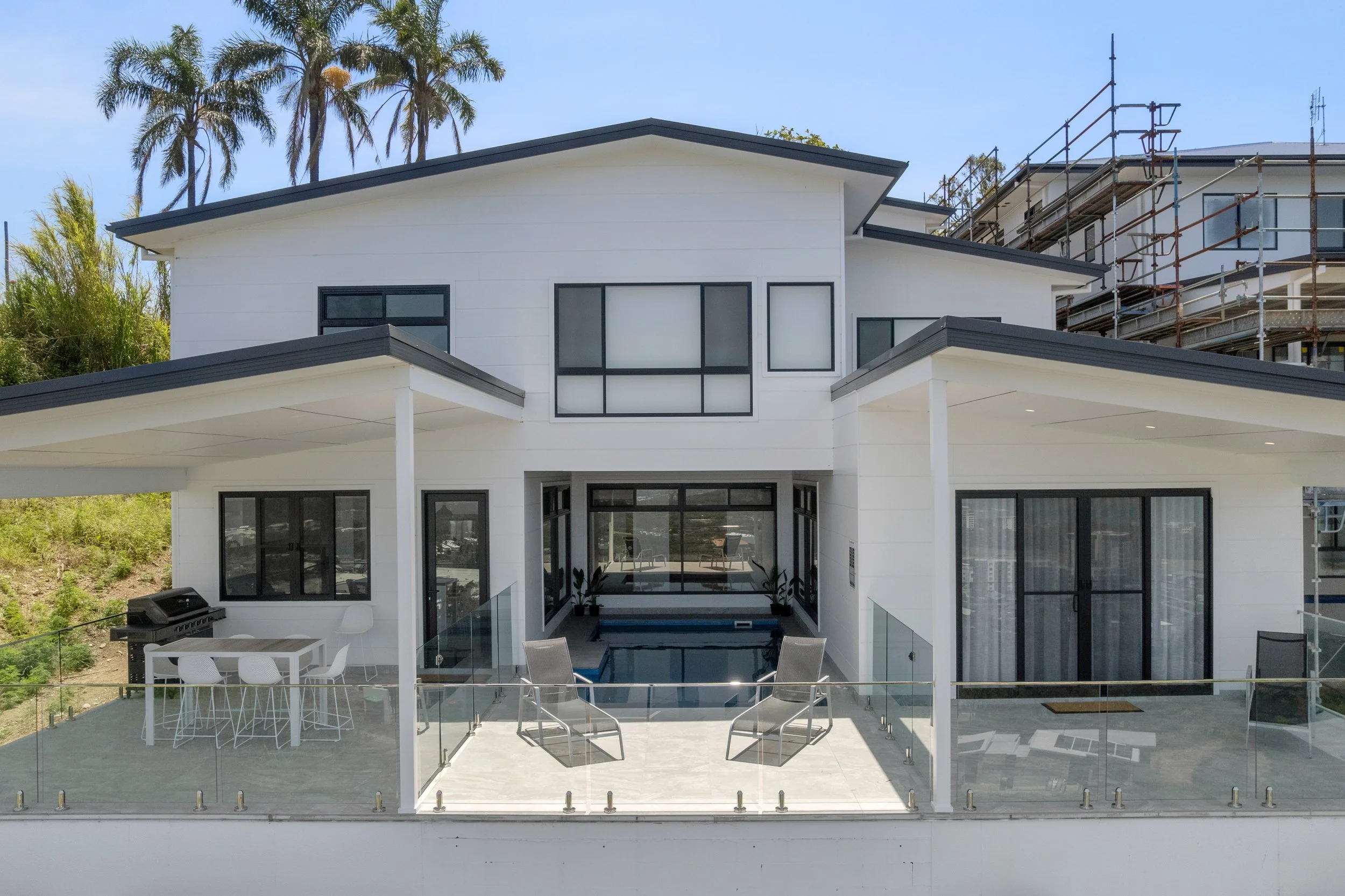 Modern white house with black window frames, a small pool, and outdoor furniture on a patio, with palm trees in the background.