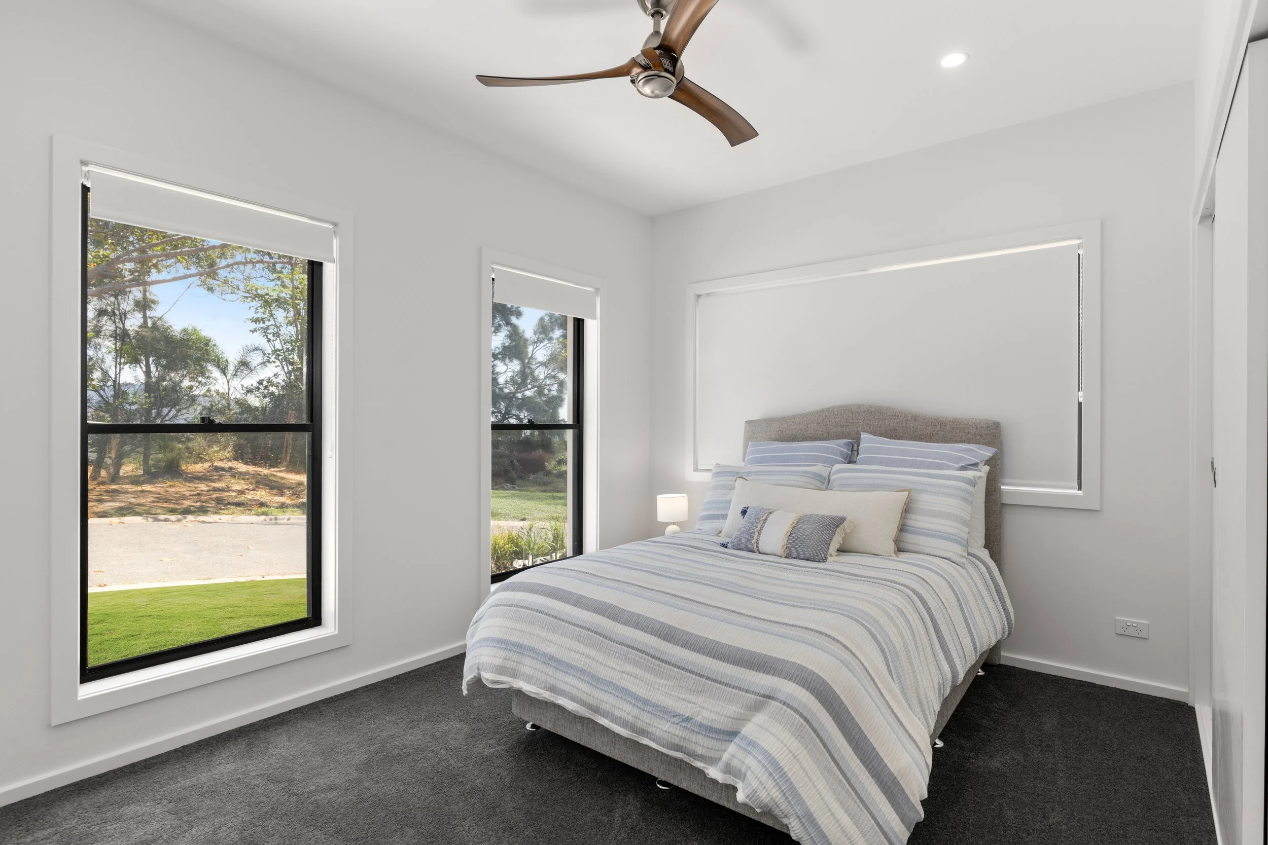 A bedroom with white walls, two large windows with black frames, a double bed with light gray upholstered headboard, striped bedspread and pillows in shades of blue, white, and beige, a small nightstand with a white lamp, ceiling fan with wooden blades, and a window with a blind on the wall above the headboard.