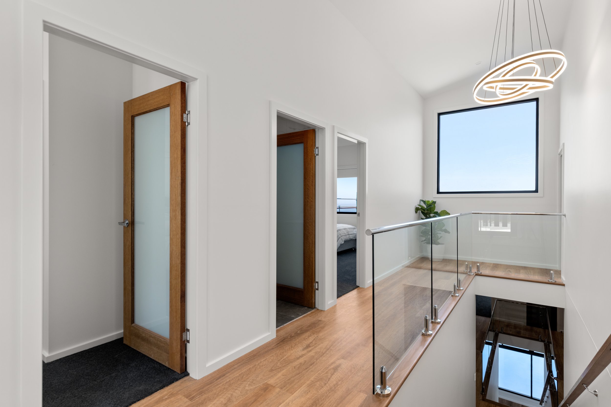 Modern indoor stair landing with white walls, wooden floors, glass railing, and contemporary ceiling light fixture. Two doors with frosted glass panels and a bedroom with a large window and potted plant visible.