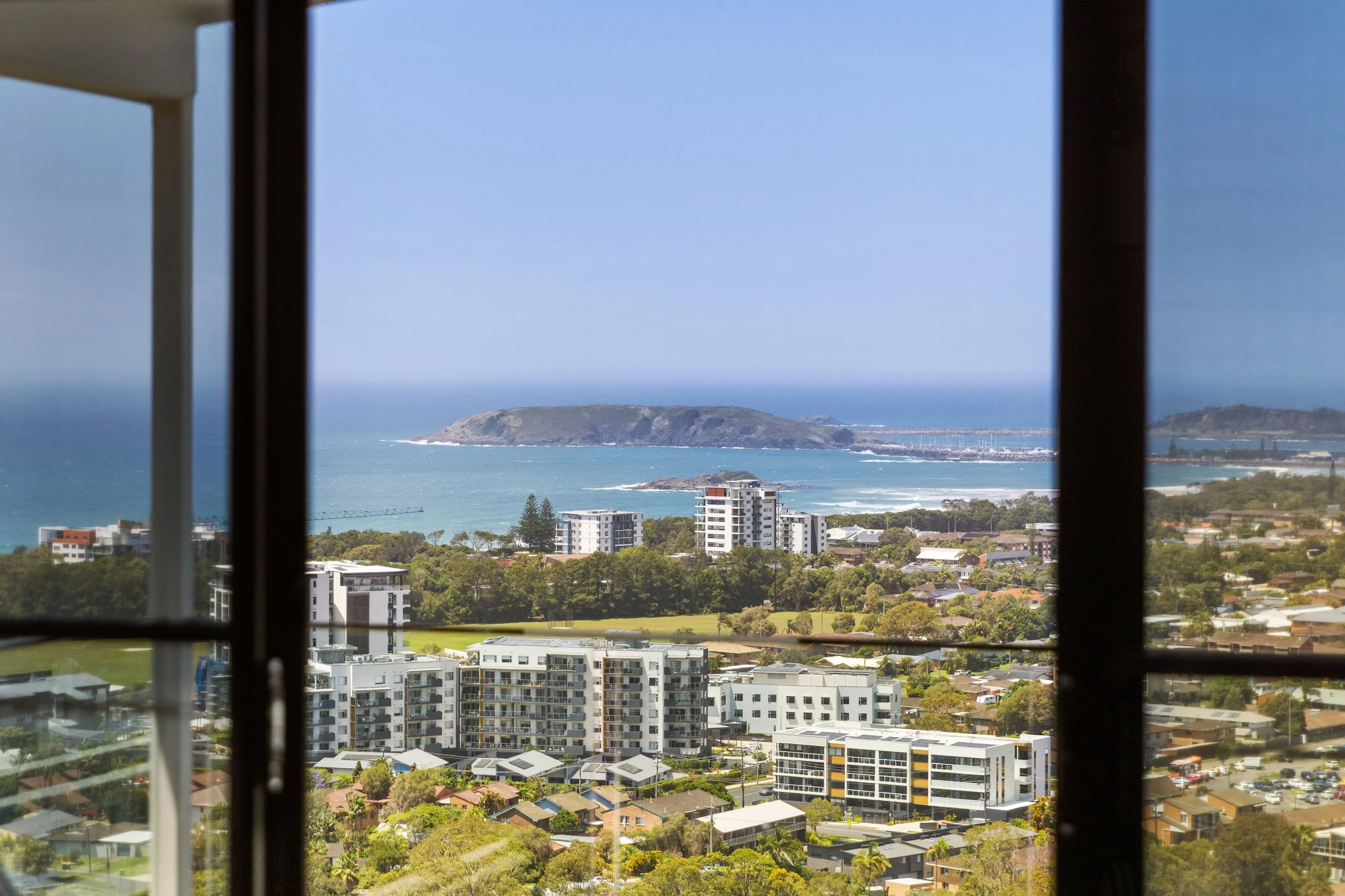 View of a coastal city seen through a window, with buildings, trees, the ocean, and an island in the distance.