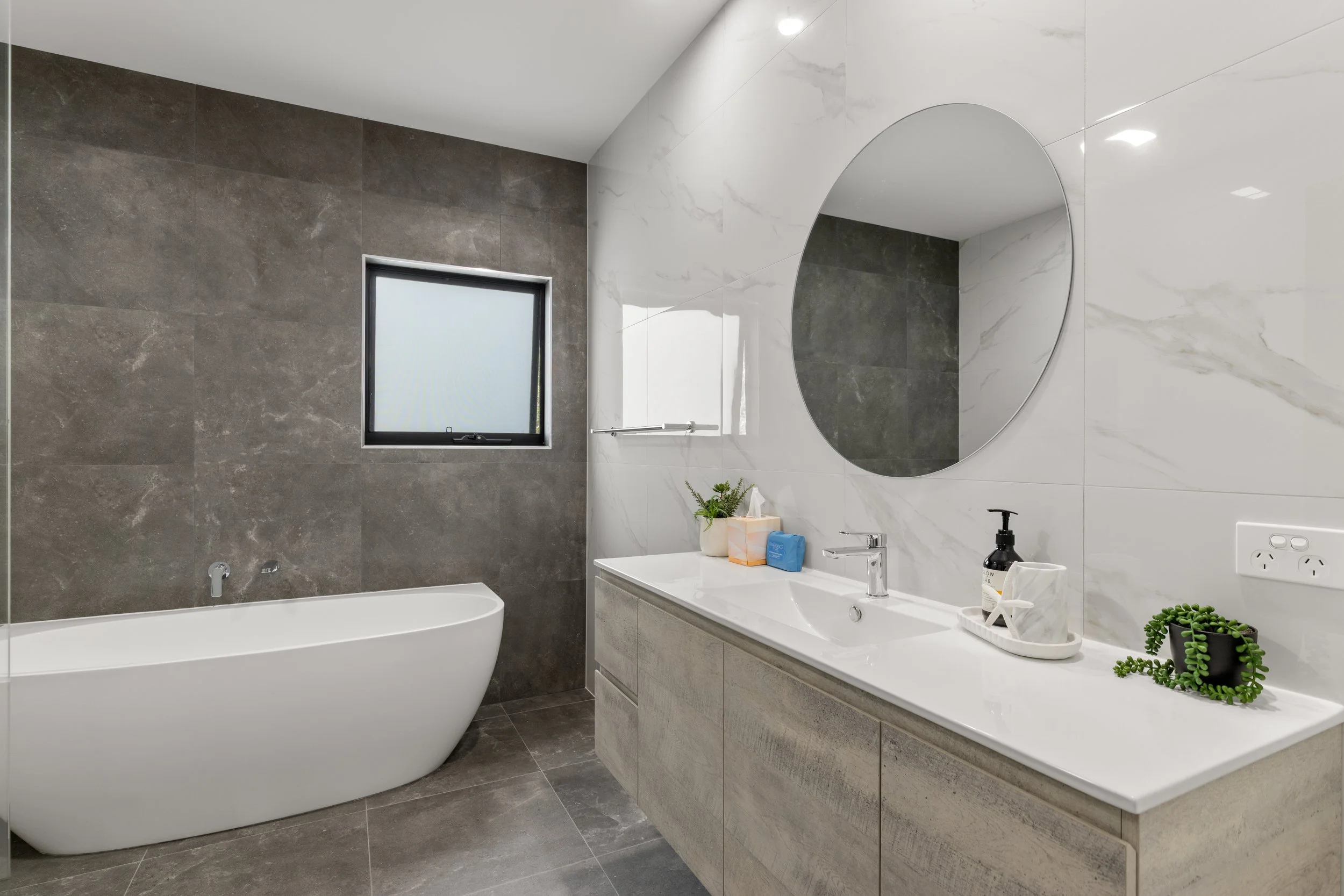 Modern bathroom with a white freestanding bathtub, a vanity with a sink, a round mirror, and decorative plants.