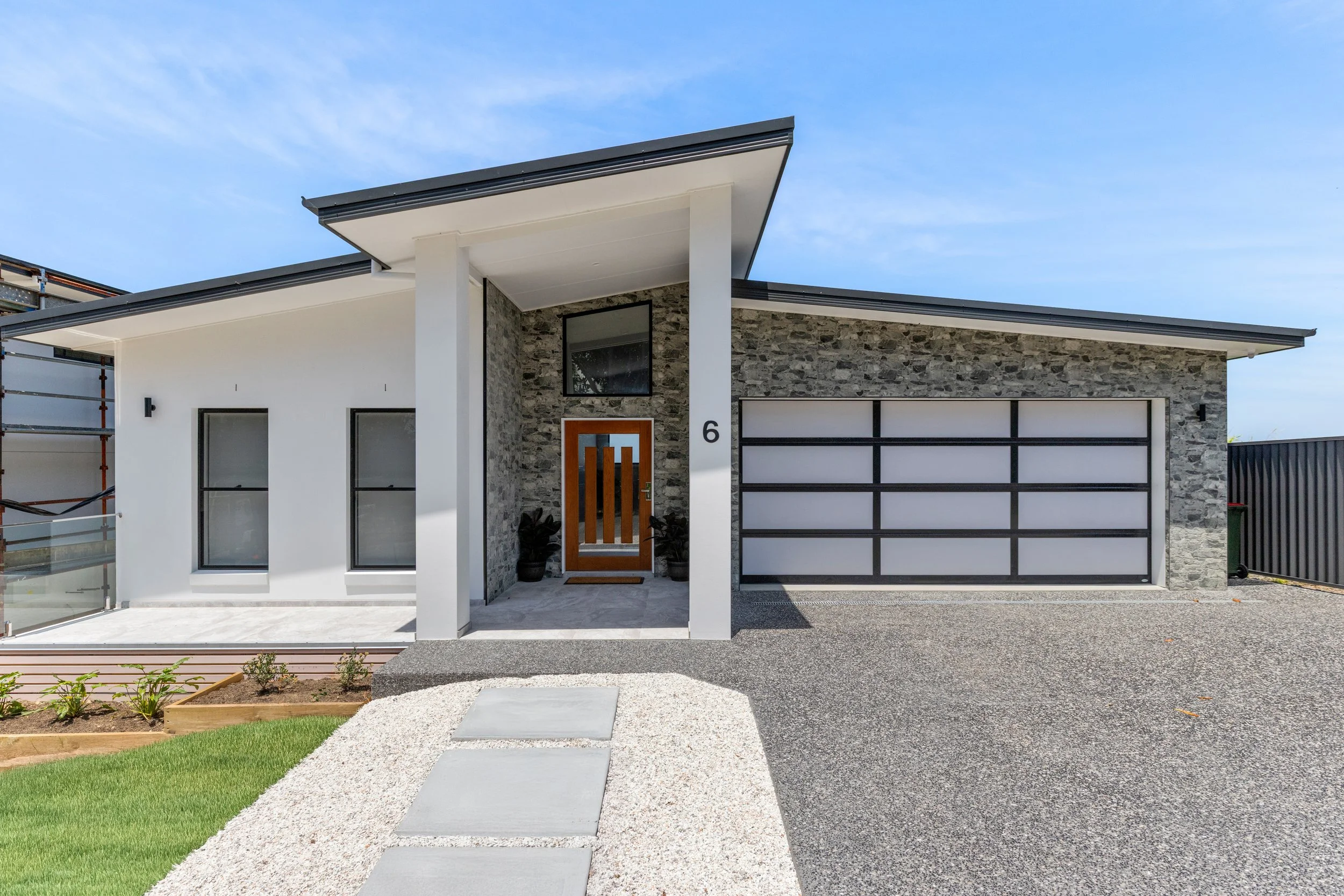 Modern house with a stone and white exterior, a wooden front door, and a large garage door, with a pathway leading to the entrance and a lawn on the side.