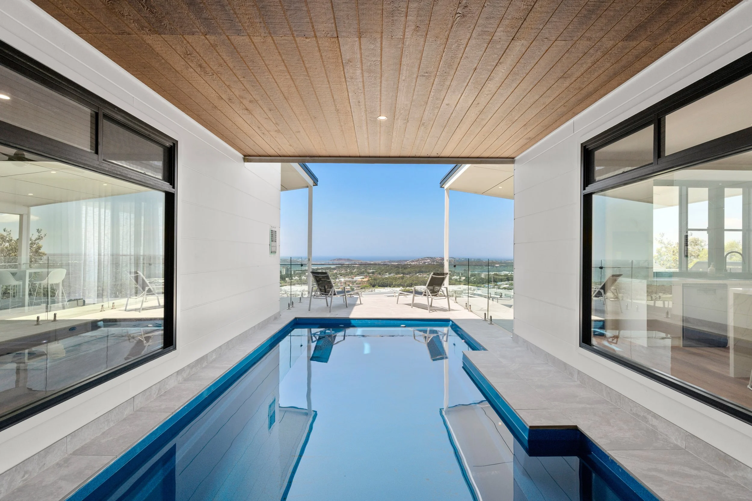 Indoor pool area with large windows, view of the outdoor balcony with chairs, and a scenic city view in the distance.