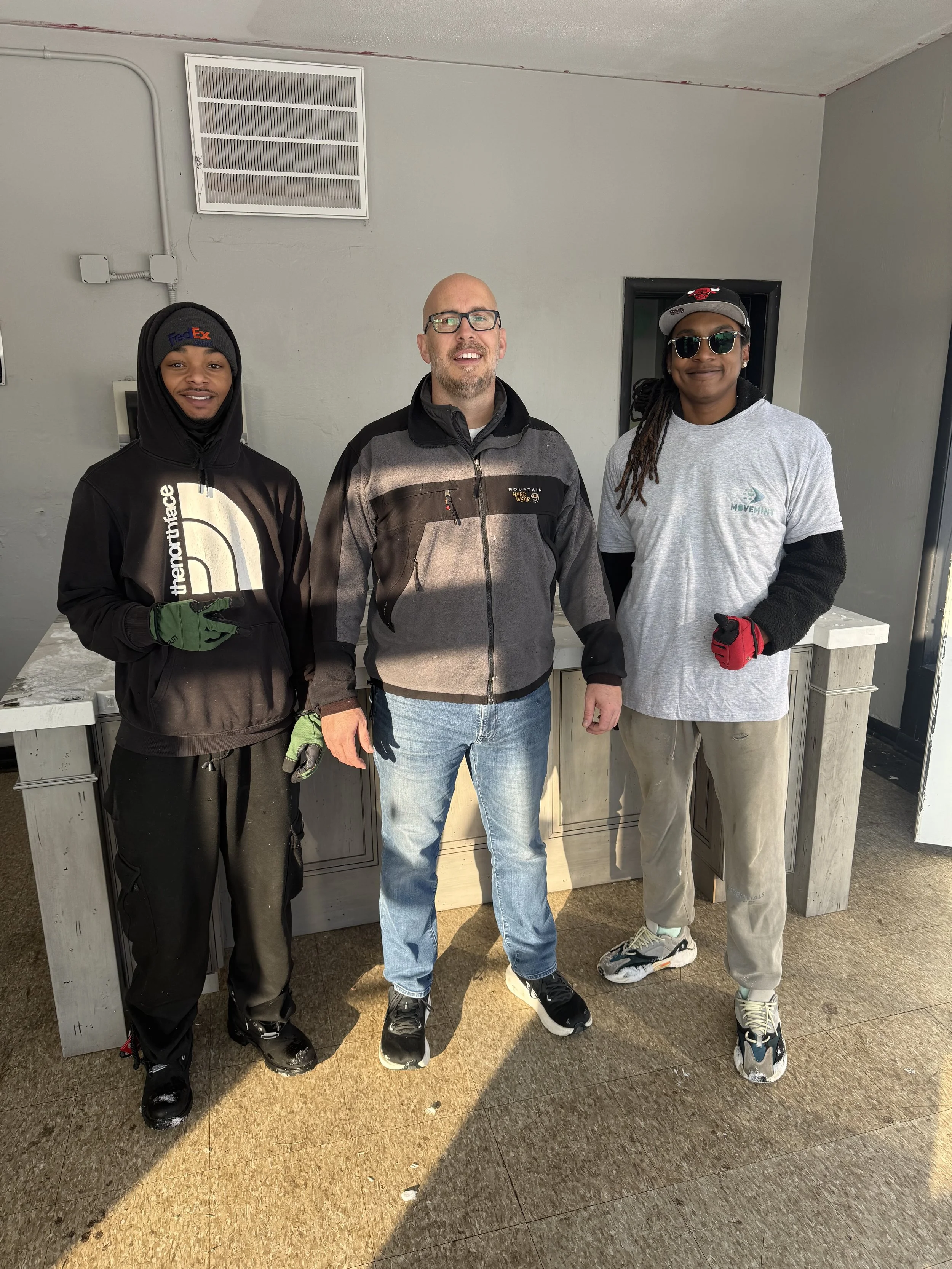 Three men standing indoors against a gray wall, smiling at the camera. They are dressed in casual and work clothing, with the man on the left wearing a black hoodie, the man in the center wearing a gray jacket, and the man on the right wearing a gray t-shirt with sunglasses.