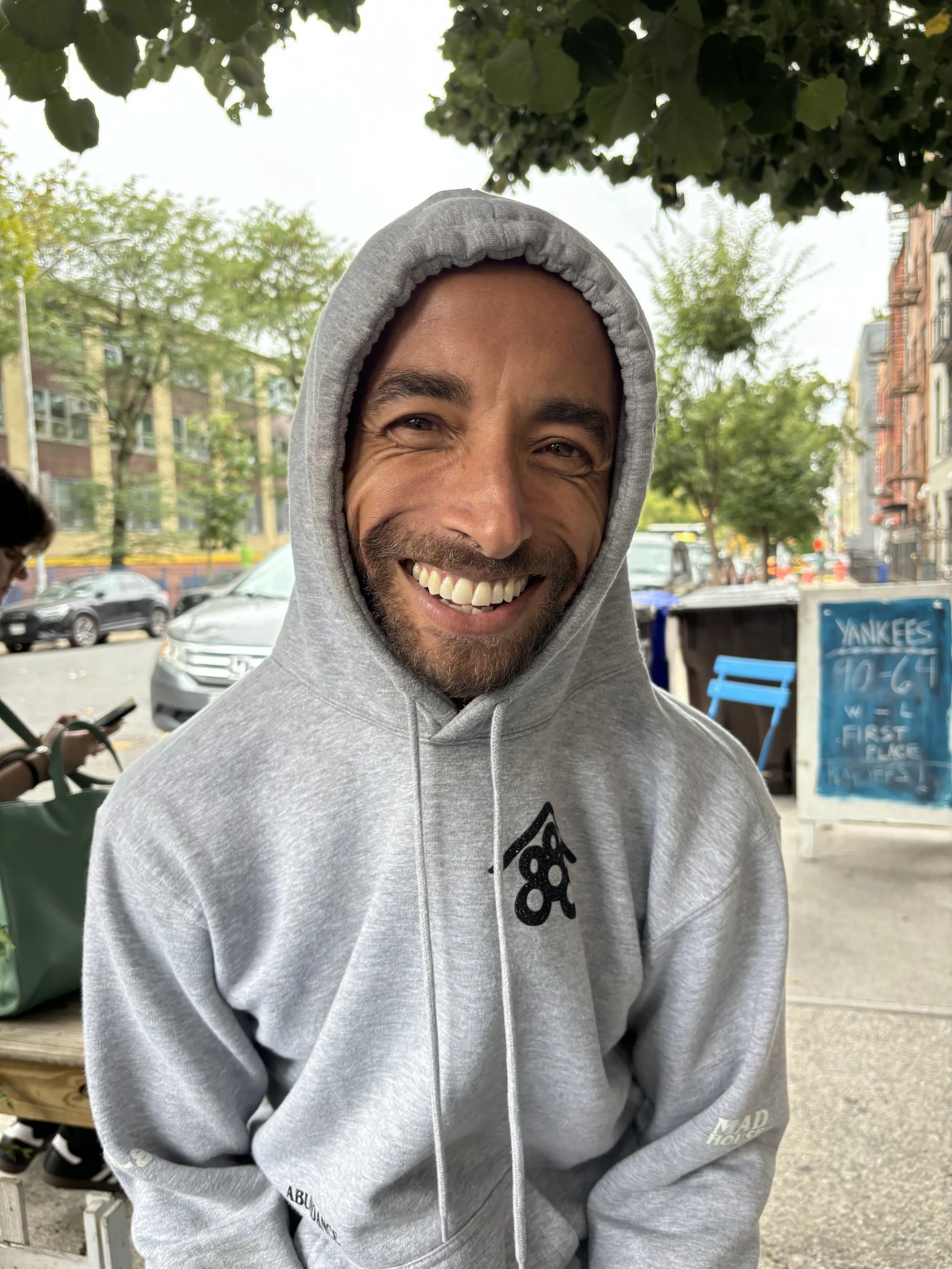 Smiling man in a gray hoodie with the hood up, outdoors on a city street with trees and cars in the background.