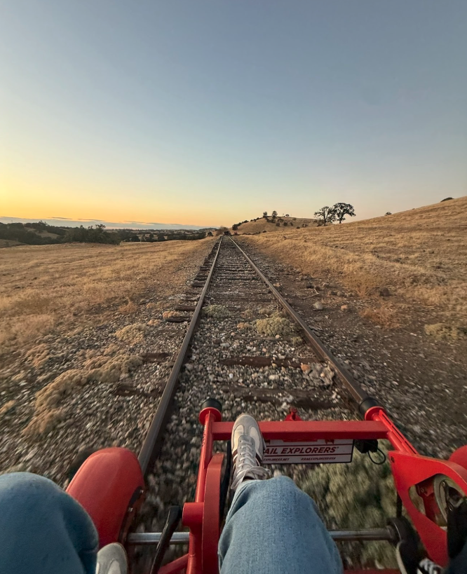 View from a red rail bike on train tracks through open fields during sunset, with a person wearing sneakers and jeans sitting in the bike.