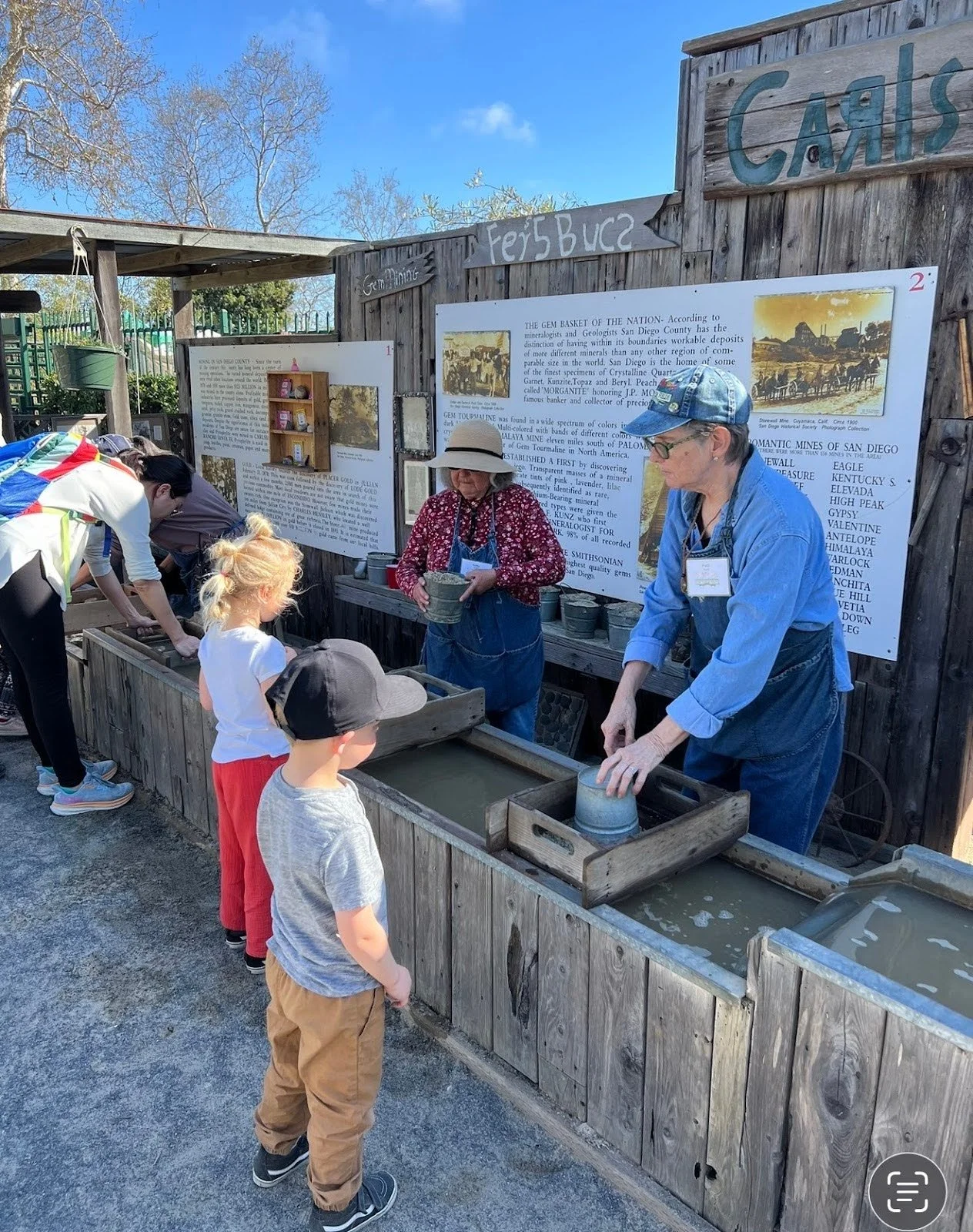 Children and adults participating in a gem crafting activity at an outdoor exhibit with informational signs about mining and geology.