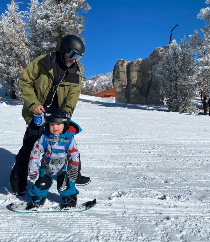 A man and a young child in winter clothing and helmets look at a snowboard on a snowy slope with snow-covered trees and a large rock formation in the background under a clear blue sky.