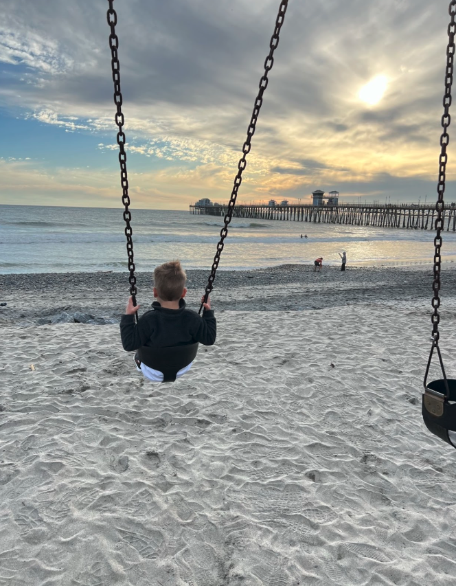 A young child sitting on a swing at the beach, facing the ocean, with a pier and buildings in the background during sunset.
