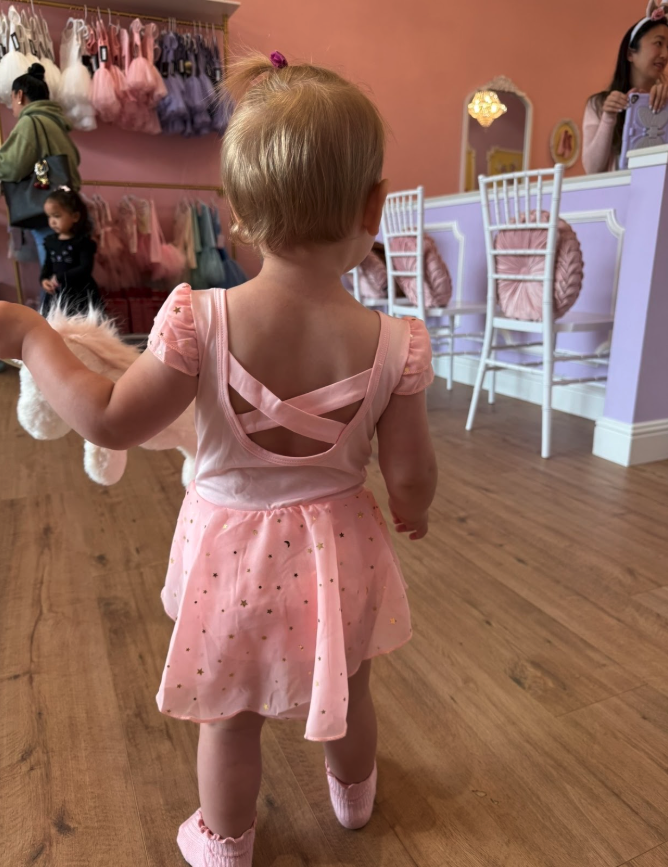 A young girl with blonde hair in a ponytail, wearing a pink dress with a crisscross back design and a sparkly skirt, holding a stuffed animal, standing inside a store with pastel-colored walls, shopping carts, and other children and adults in the bac