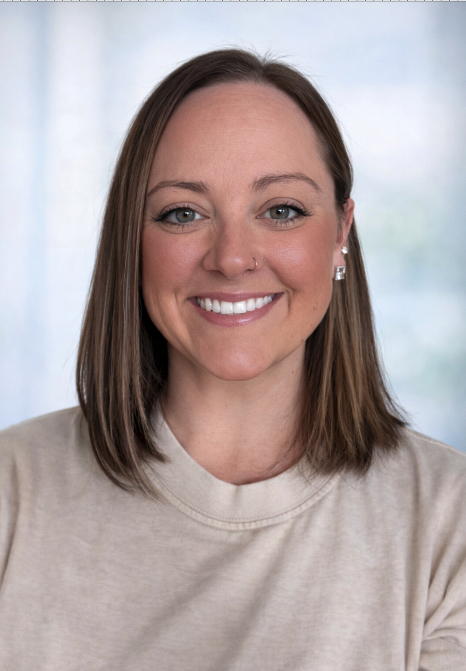 A woman with shoulder-length brown hair, blue eyes, and a nose piercing, smiling while wearing a beige t-shirt and earrings, with a blurred light-colored background.