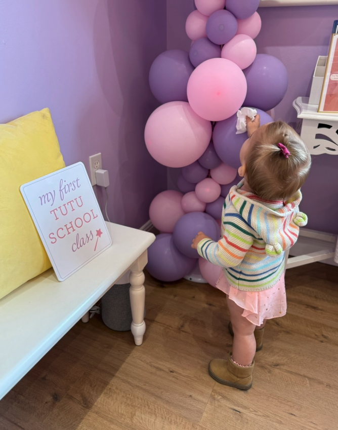 A young girl with a ponytail, wearing a striped rainbow sweater, a pink skirt, and brown boots, reaching for pink and purple balloons. There is a sign on a small white table that says 'my first TUTU school day' and a yellow pillow. Purple walls and a