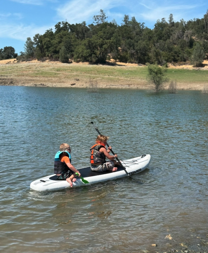 Two children on a paddleboard in a lake, wearing life jackets and one holding a paddle. The lake is surrounded by trees and a grassy bank, with a blue sky overhead.