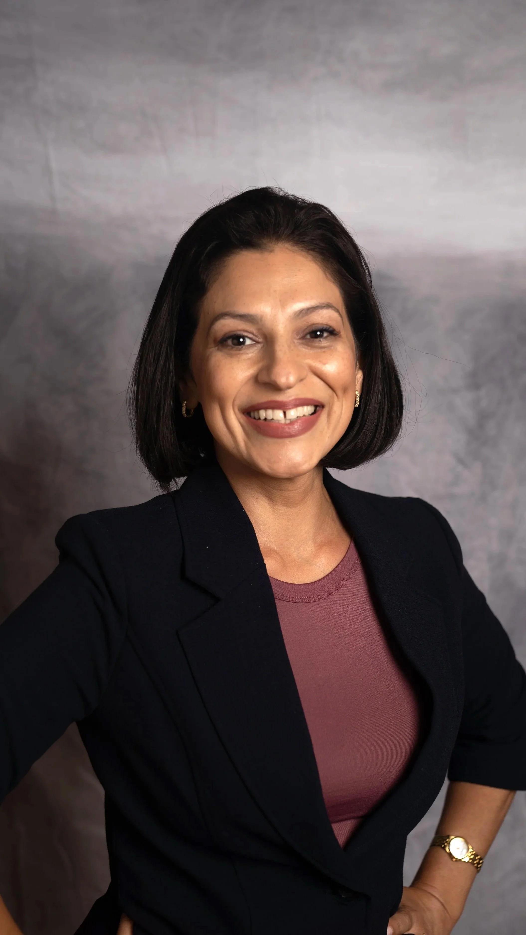 Los Angeles Board of Secretary Candidate, Noel Almario, wearing a black blazer and maroon top, smiling with her hand on her hip, against a gray textured background.