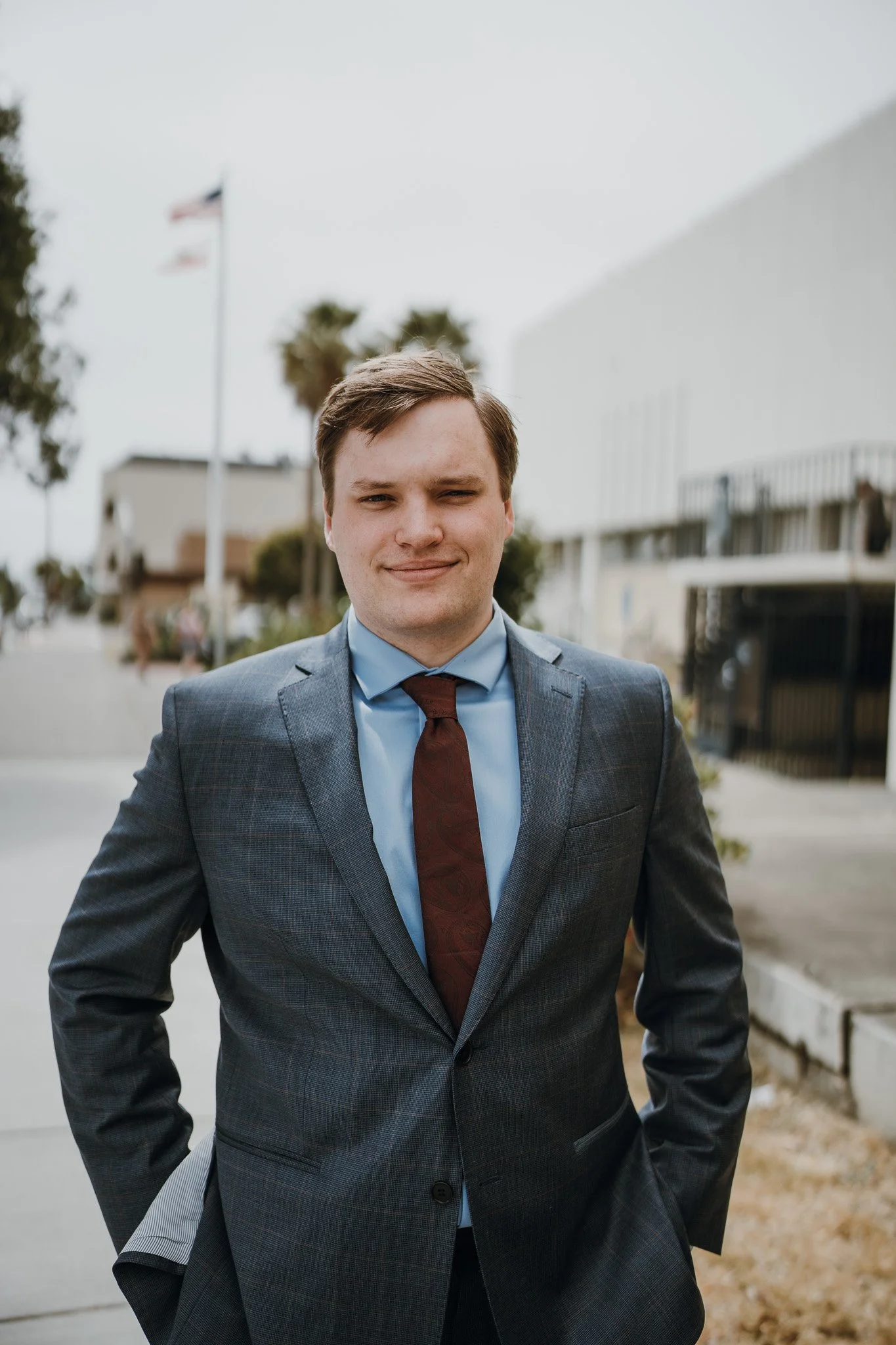 A young man in a gray plaid suit with a blue shirt and maroon tie standing outdoors, smiling with his hands in his pockets, in front of a city street scene with a building, trees, and some flags in the background in Humboldt County taken by wedding a