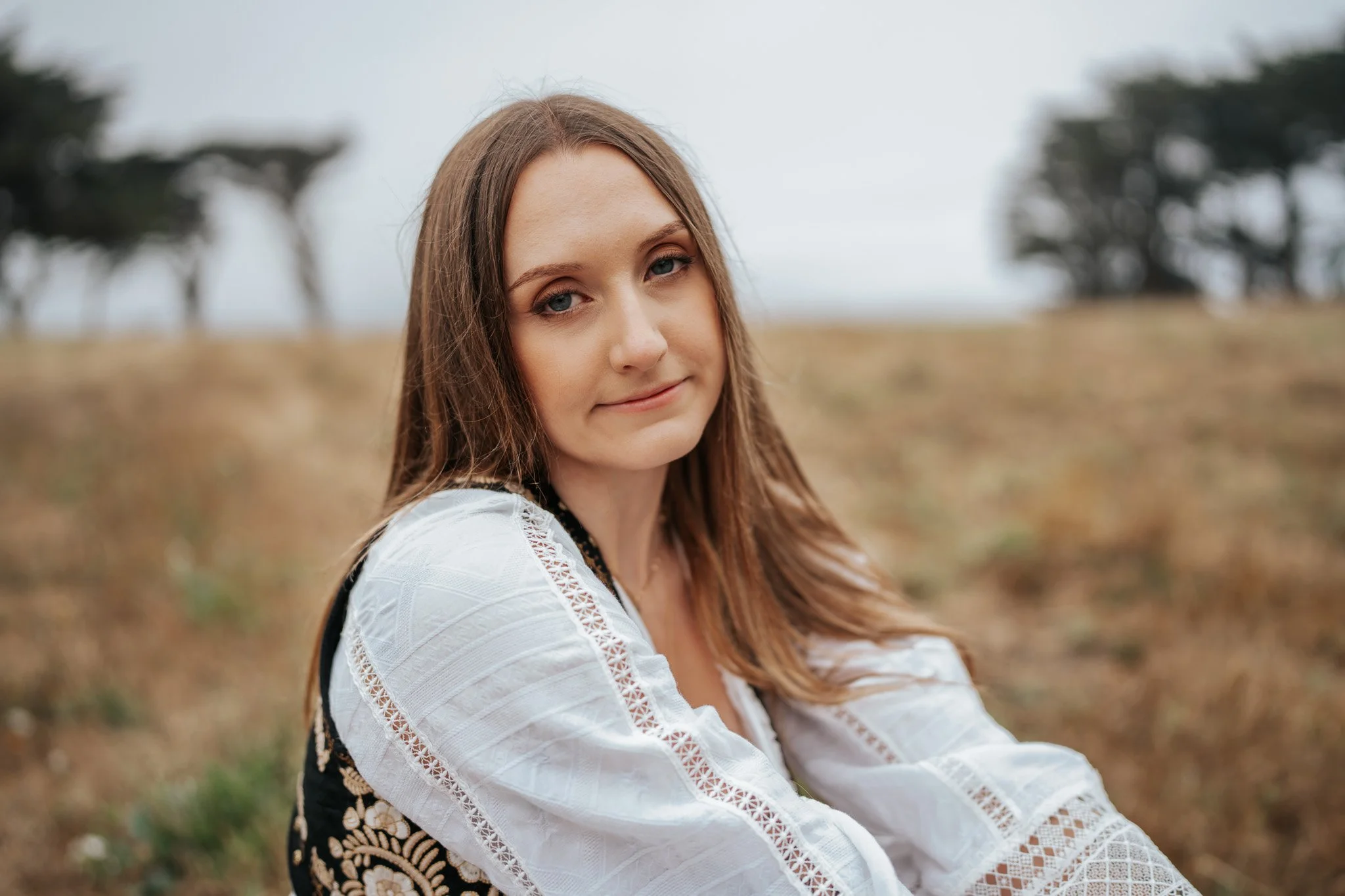Young woman with long light brown hair and light skin, smiling softly, sitting outdoors on a grassy plain with blurred trees in the background in Humboldt County taken by wedding and elopement photographer, Molly Kelly.