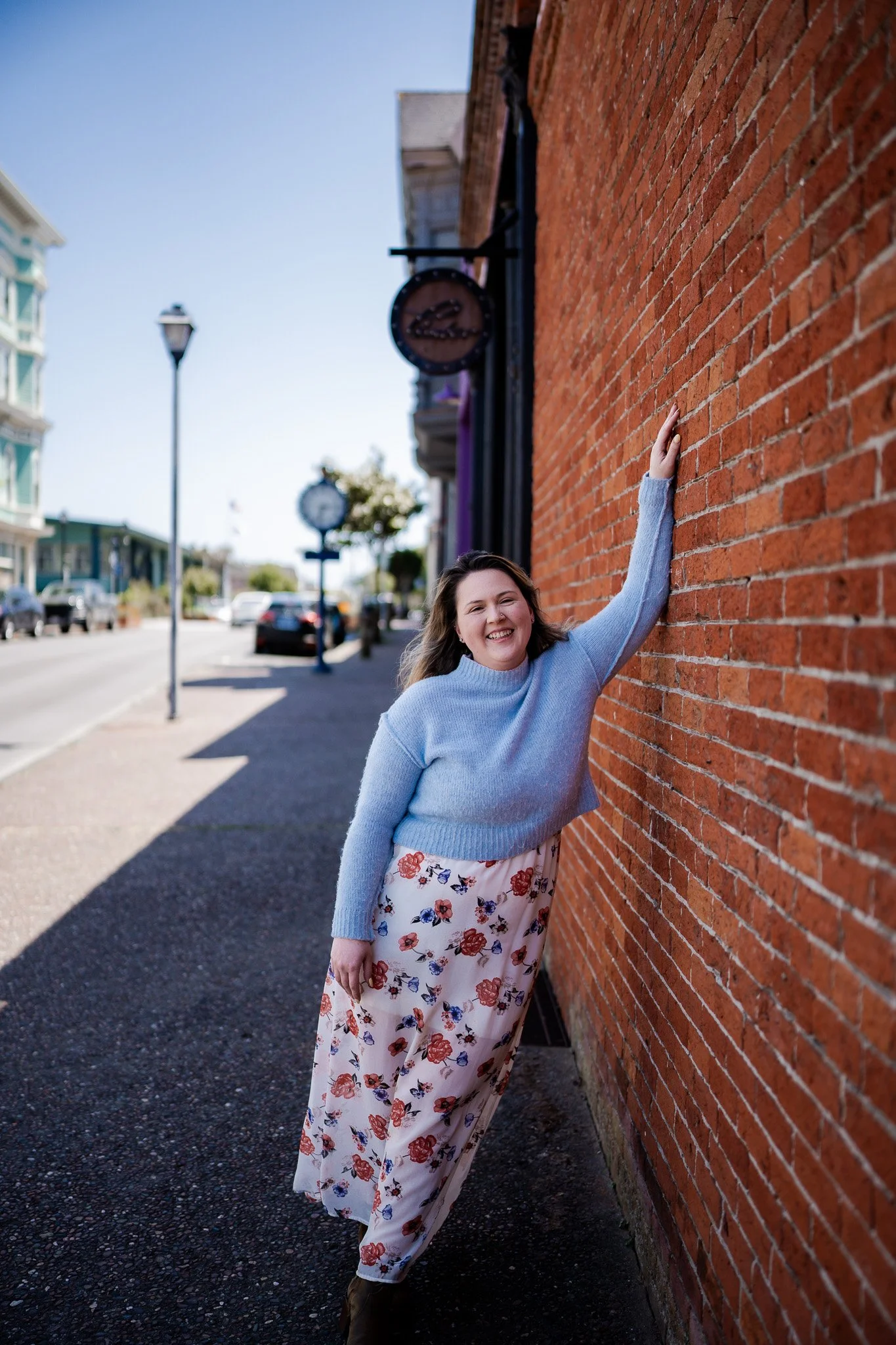 A smiling woman in a light blue sweater and floral pants standing on a sidewalk and leaning against a red brick wall in Humboldt County taken by wedding and elopement photographer, Molly Kelly.