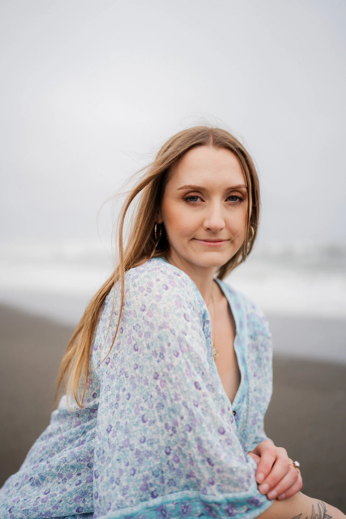 A young woman with long, light brown hair and blue eyes is sitting on a beach, wearing a light blue floral blouse, with a calm, overcast sky and ocean in the background in Humboldt County taken by wedding and elopement photographer, Molly Kelly.