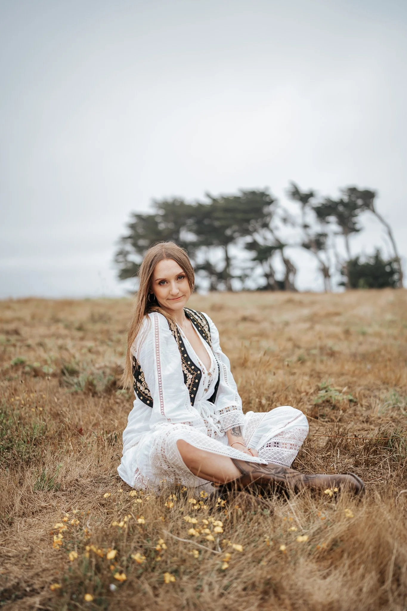A young woman with long brown hair sitting on a grassy field, wearing a white embroidered dress and boots, with trees in the background under an overcast sky in Humboldt County taken by wedding and elopement photographer, Molly Kelly.