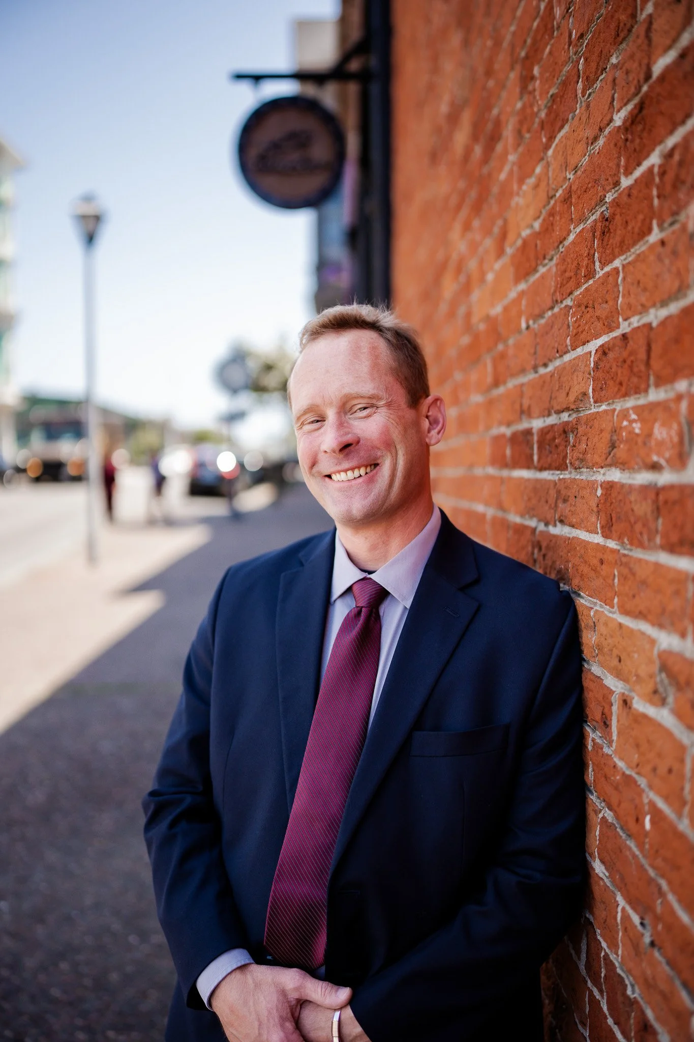 A man in a navy suit and maroon tie smiling while leaning against a brick wall on an outdoor sidewalk, with traffic and street signs in the background on a sunny day in Humboldt County taken by wedding and elopement photographer, Molly Kelly.