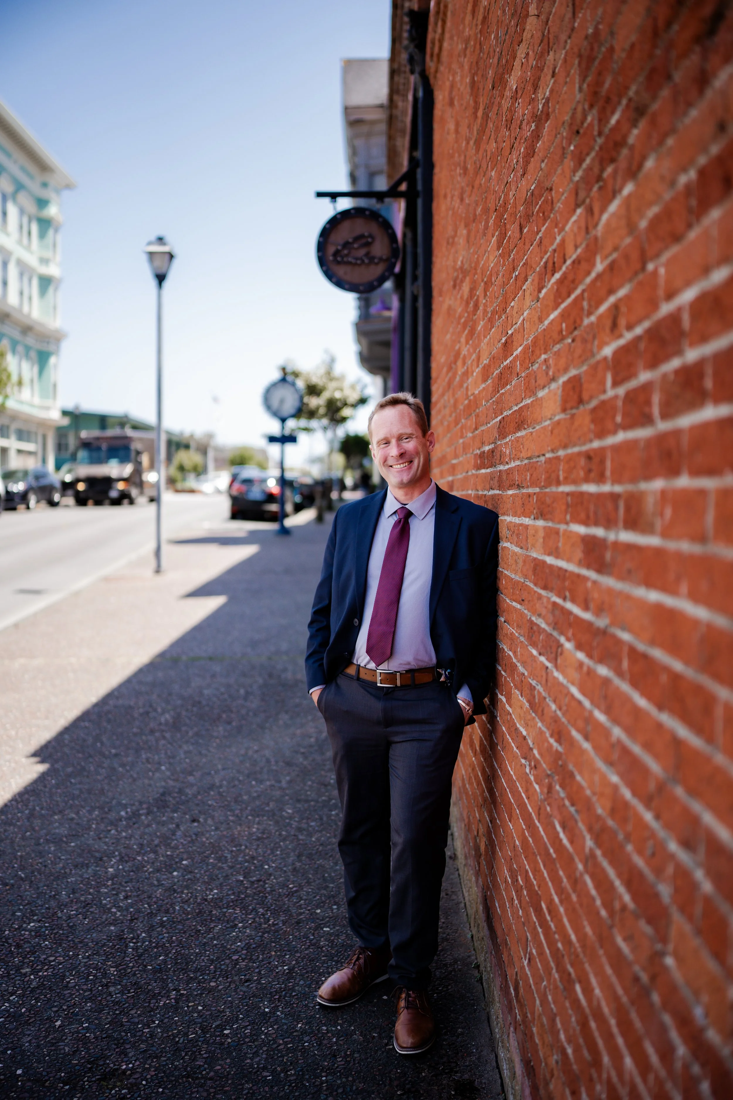 A man in a suit smiling and leaning against a brick wall on a sidewalk in a downtown area in Humboldt County taken by wedding and elopement photographer, Molly Kelly.