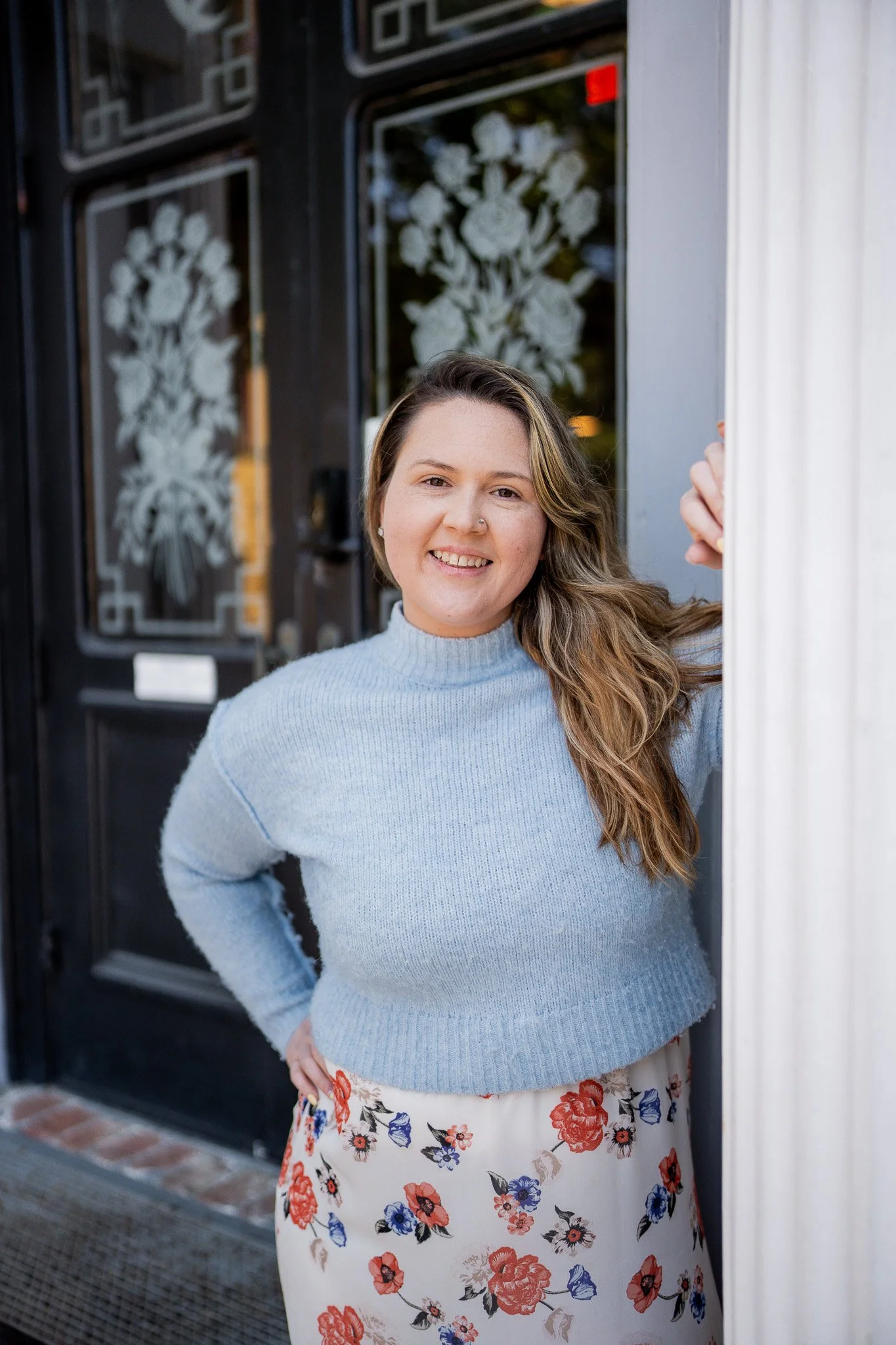 A woman with long, wavy hair smiling and standing outside a restaurant entrance, wearing a light blue sweater and a floral skirt in Humboldt County taken by wedding and elopement photographer, Molly Kelly.