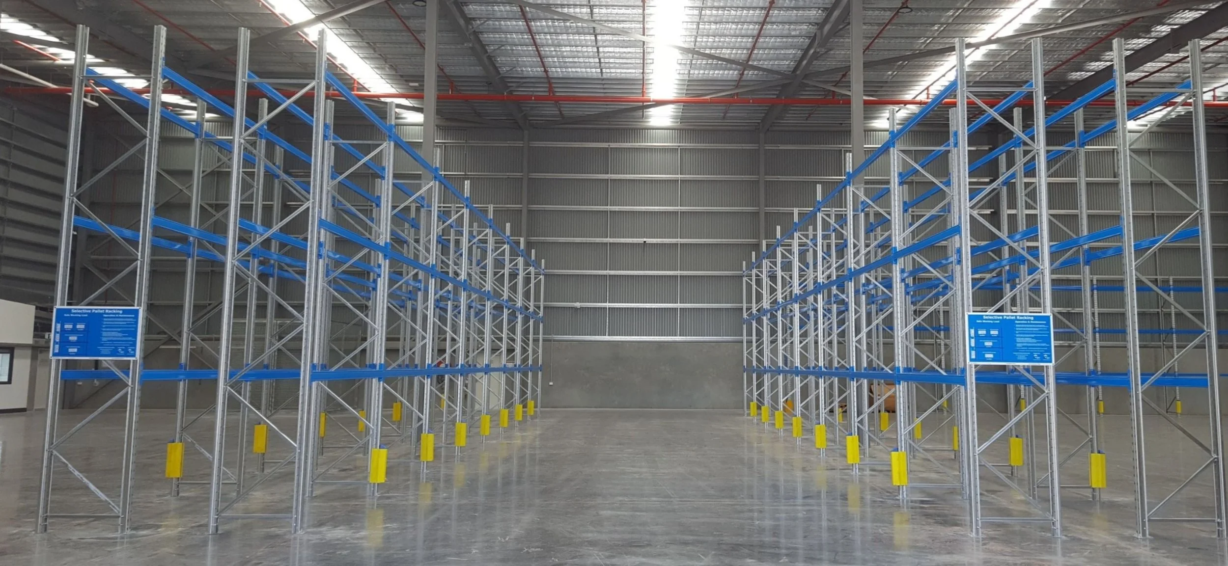 Empty warehouse with blue and silver metal pallet racks and yellow safety bumpers, labeled with blue informational signs.