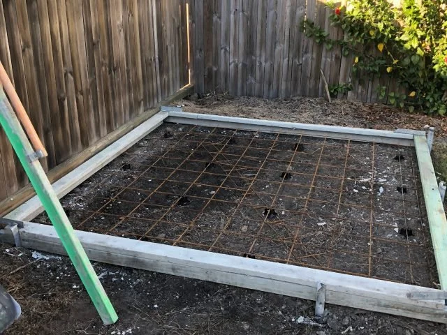 A backyard garden bed frame with a wire grid installed, surrounded by a wooden fence and a green garden tool leaning against the fence.