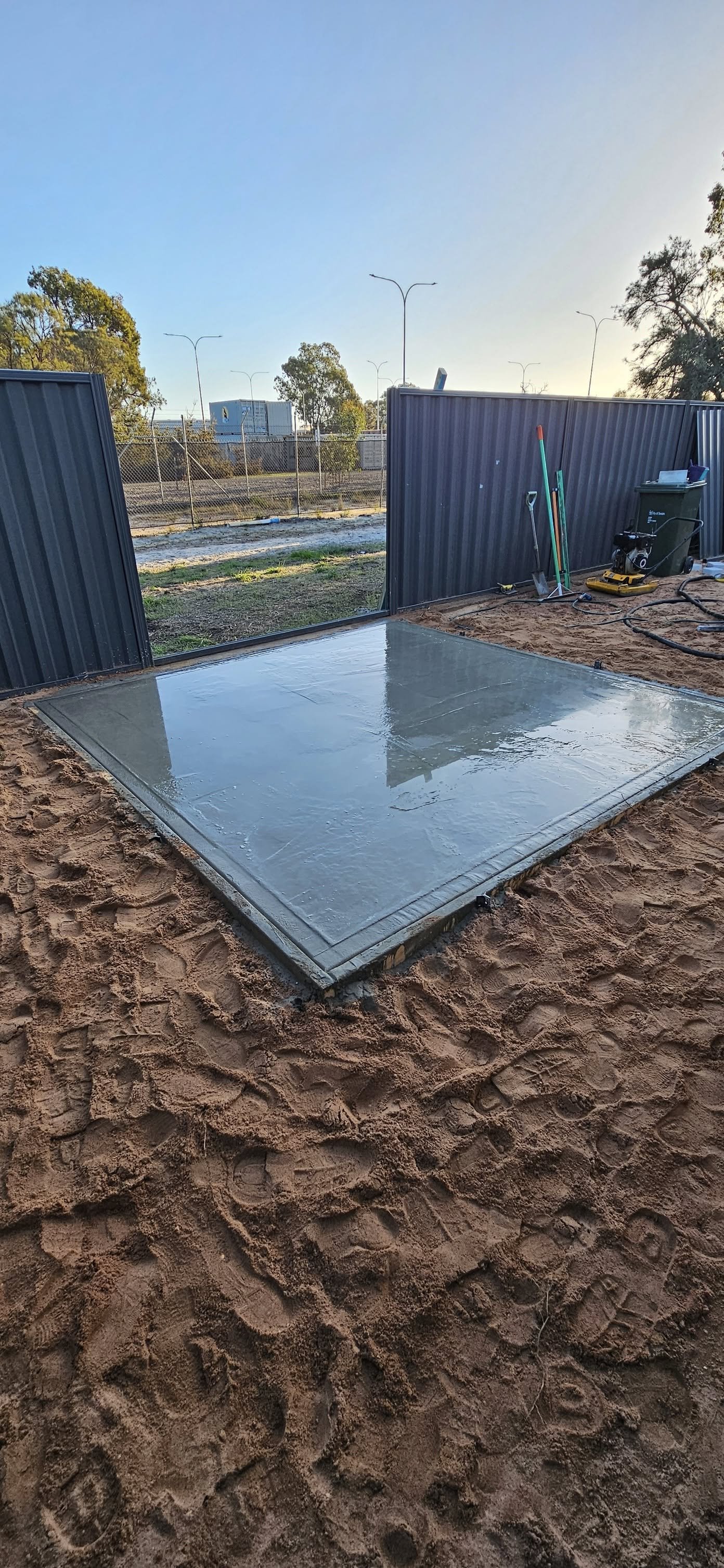 Clean cement slab in a sandy outdoor construction area, with tools and a grey fence, under a clear blue sky.