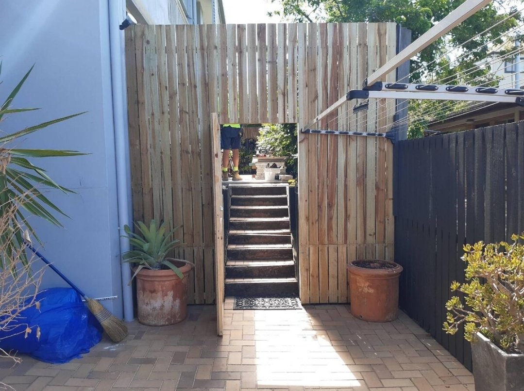 A fenced backyard area with a small staircase leading up to a gate. Potted plants and gardening tools are visible on the patio.