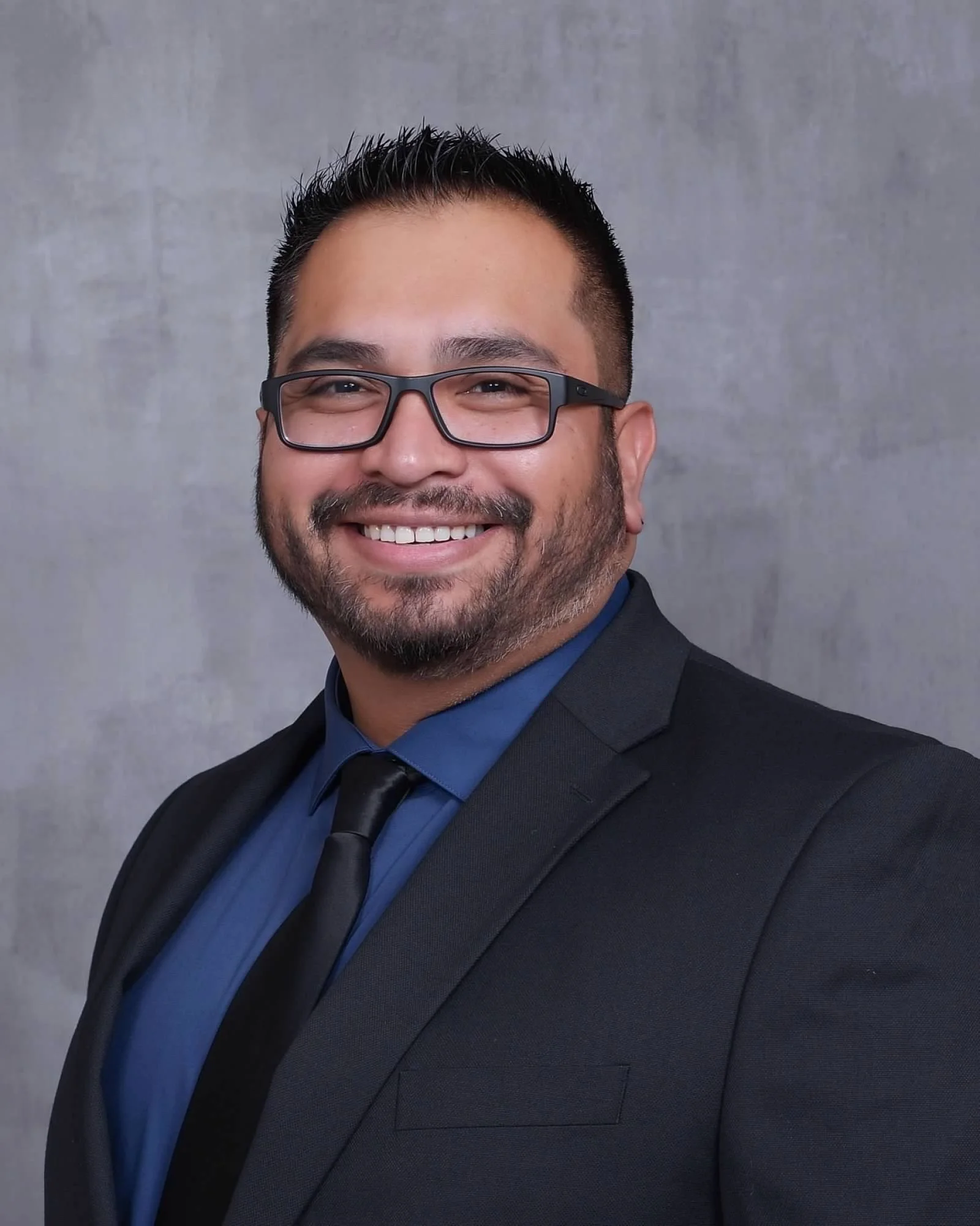 Kevin Cummings. Close-up of a man with glasses, smiling, wearing a suit and tie, against a plain gray background.
