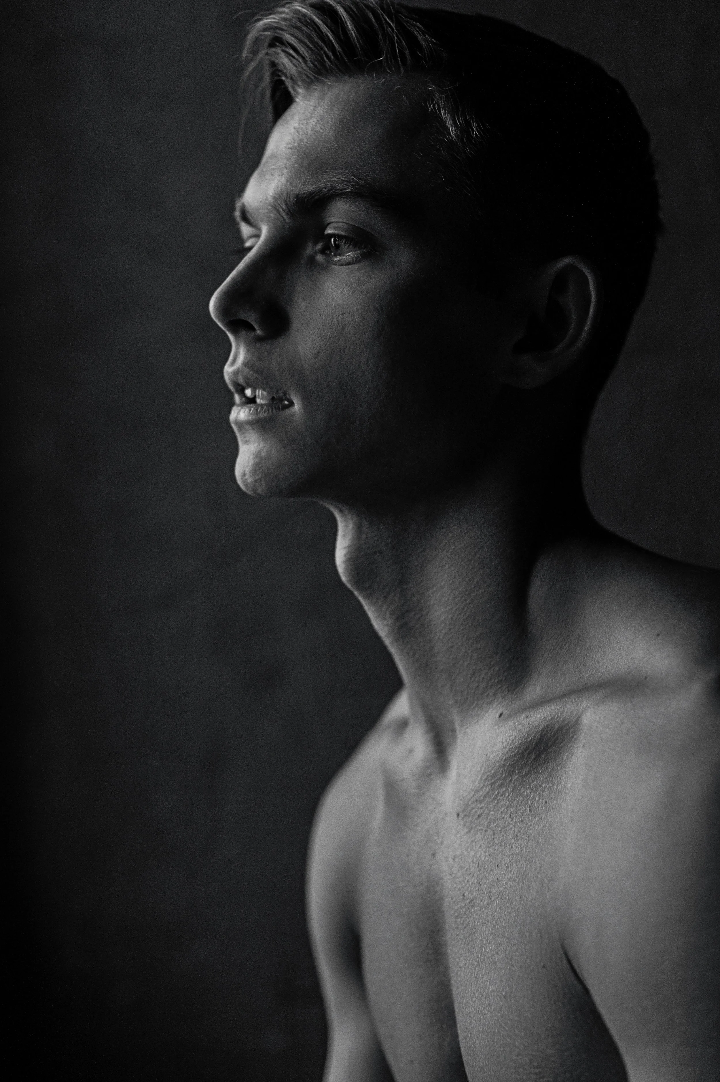 Black and white portrait of a young shirtless man with short, styled hair, looking to the side against a dark background.