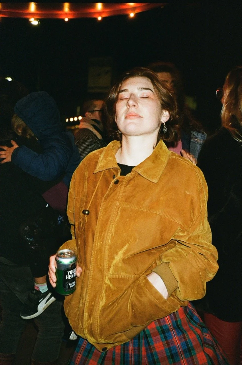 A woman with short brown hair wearing a mustard yellow jacket and a tartan skirt, holding a can of beer, standing with her eyes closed at a social gathering indoors.
