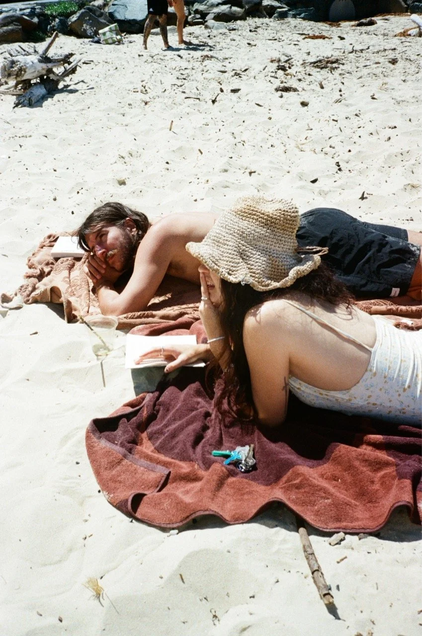 A man and woman lying on a beach towel on sand, talking, with the man resting his head on his hand and the woman wearing a straw hat.