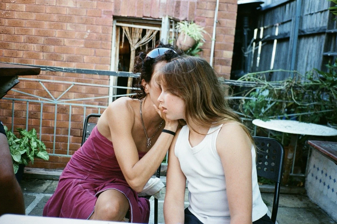 Two women sitting close together outside, one whispering into the other's ear with eyes closed, in a garden or patio area.
