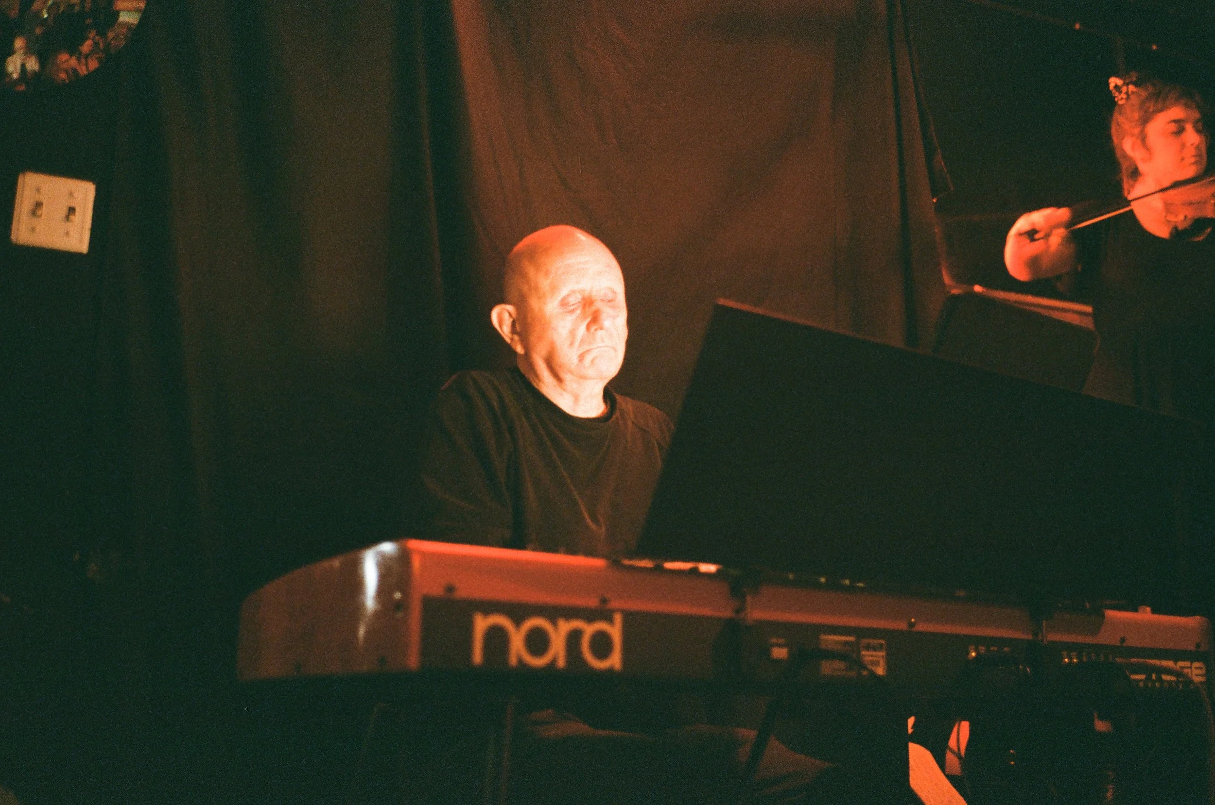 An older man with a shaved head playing a Nord keyboard on stage, with a woman in the background holding a violin.