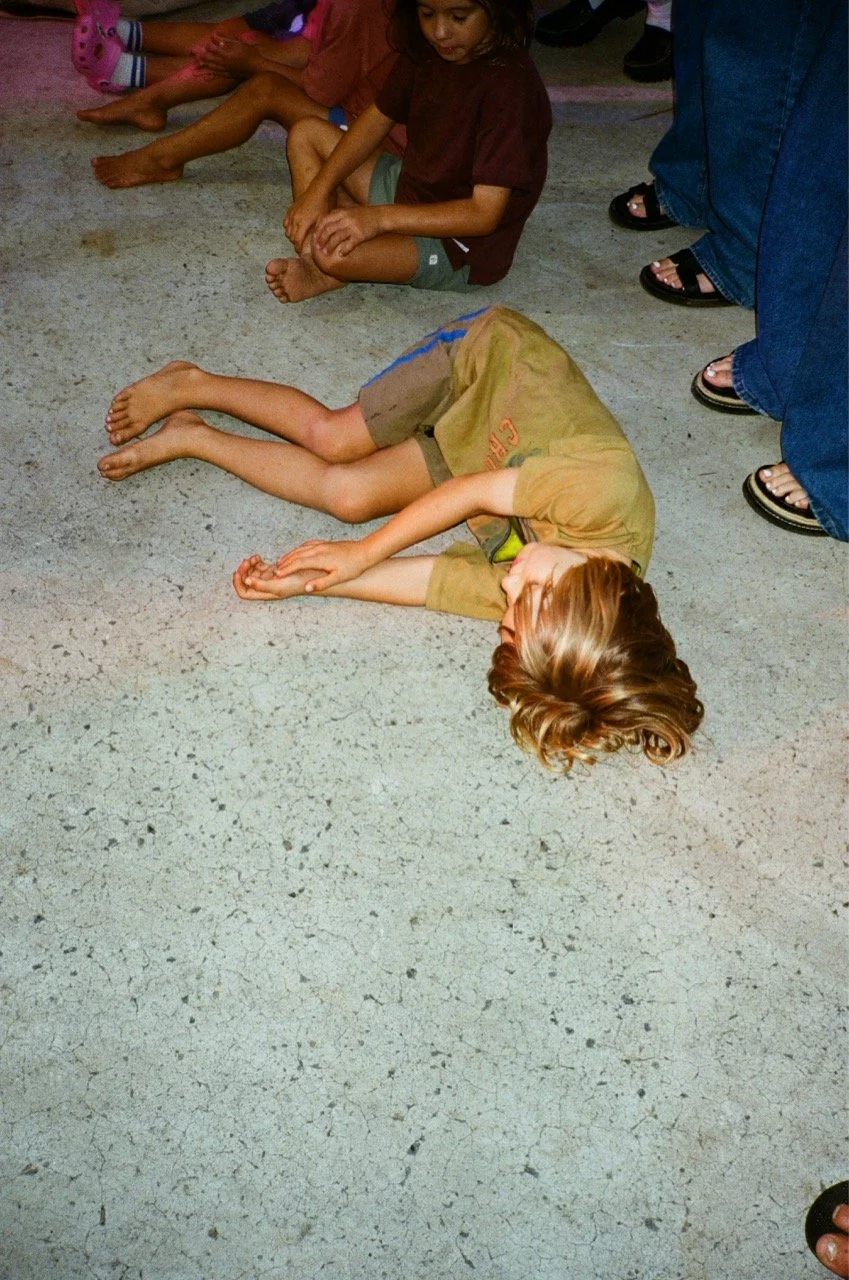 A young child is lying face down on a concrete floor, with others sitting nearby. The child appears to be resting or possibly in distress, and adults are standing around.