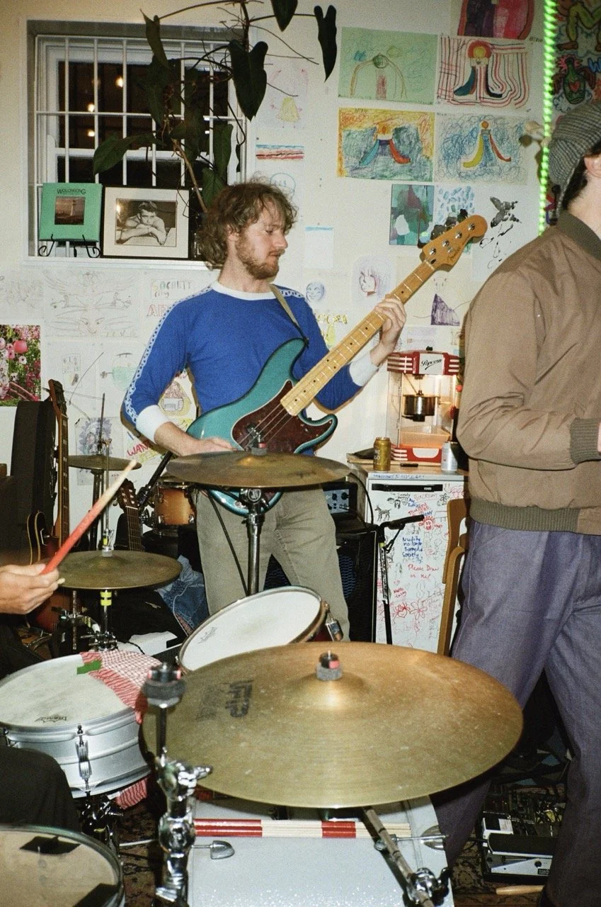 A man with curly hair and a beard plays a teal electric bass guitar at a small indoor musical gathering, surrounded by drums and other instruments, with children's drawings on the wall behind him.