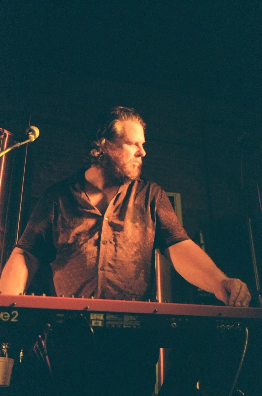 A man with a beard and long hair playing a keyboard on stage under dim lighting.
