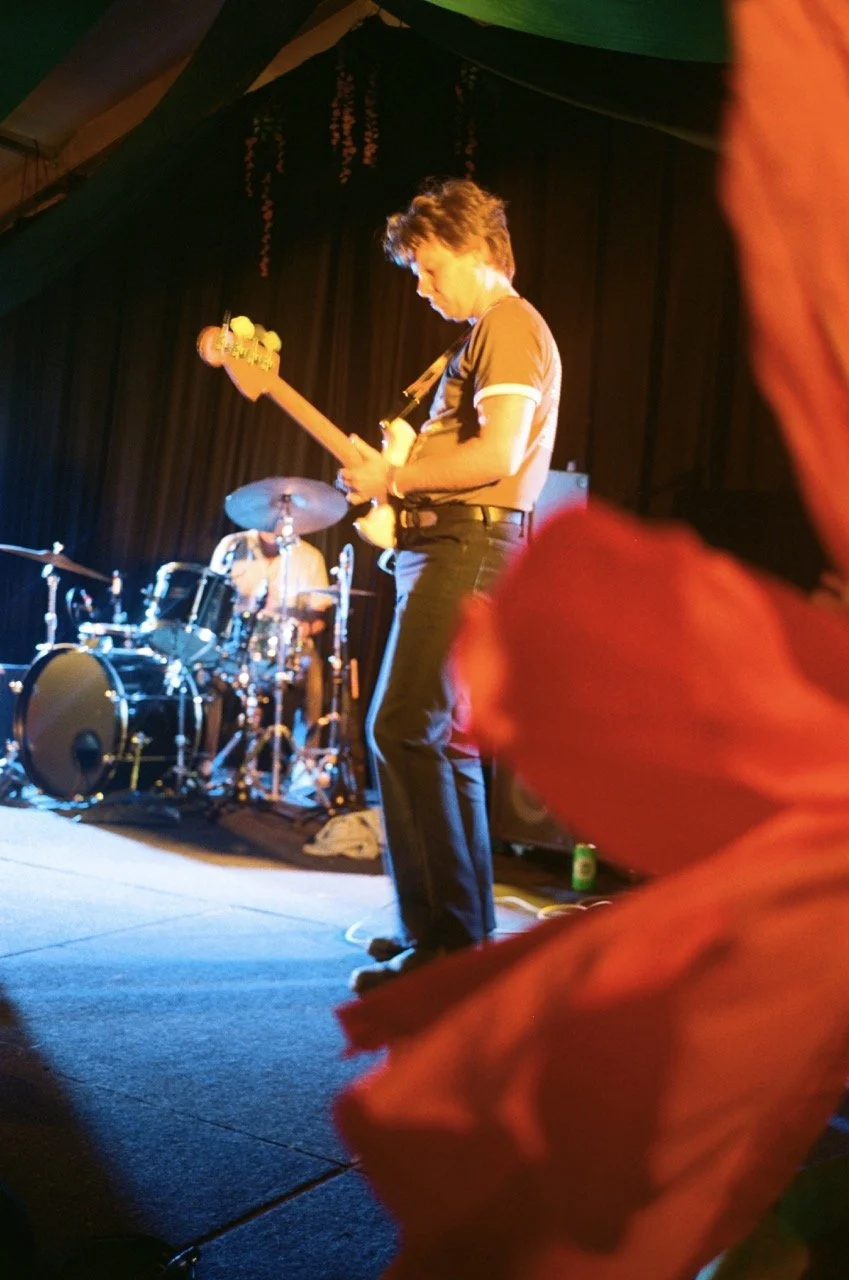 A male musician playing an electric guitar on stage with a drummer behind him. The stage is dimly lit with warm colors, and a black curtain backdrop is visible.