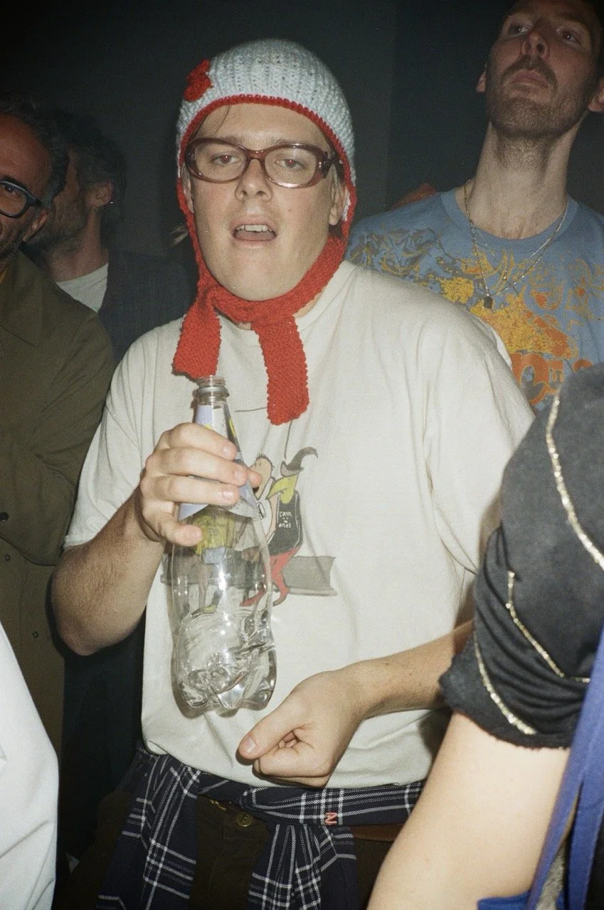 A young man wearing glasses, a knit hat with a red flower, and a t-shirt with a cartoon graphic, holding a plastic bottle, at a social gathering.