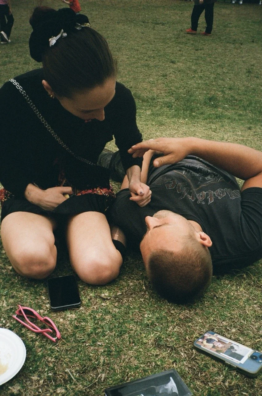 A woman kneeling on grass, holding and comforting a man lying on the ground, with a phone and sunglasses nearby.