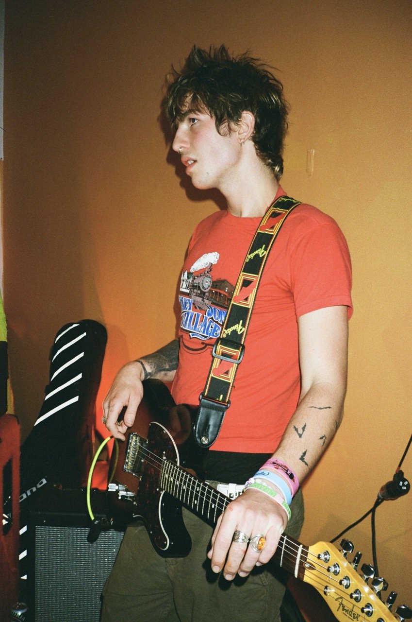Young man with messy brown hair wearing a red graphic T-shirt and multiple bracelets playing an electric guitar.