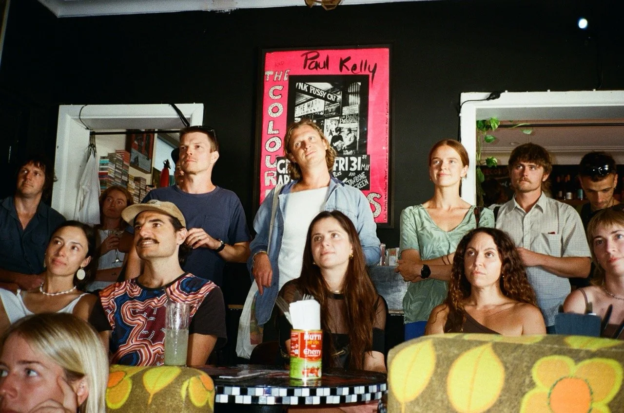 A group of people gathered indoors, some sitting and some standing, attentively watching something. There is a black wall with a vibrant pink poster behind them, featuring a black and white photo and text.