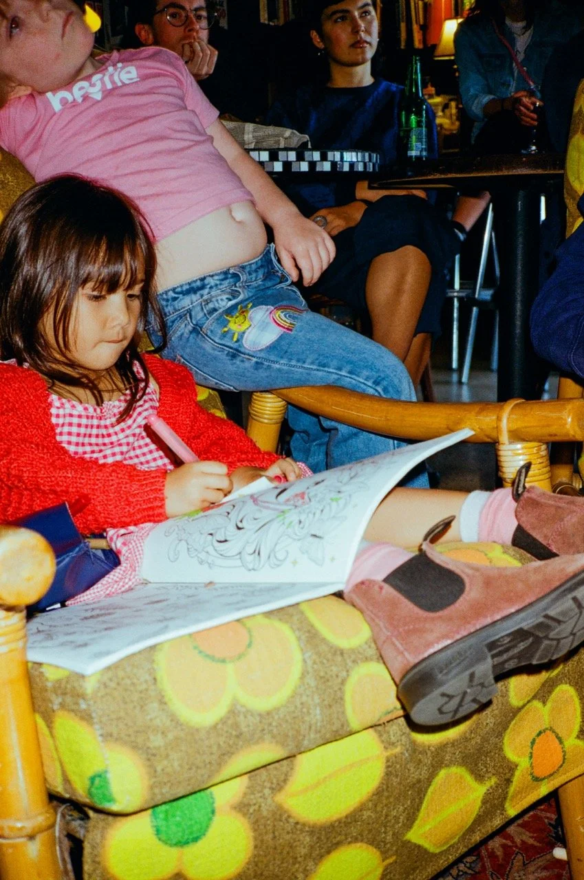 A young girl sitting on a floral-patterned chair, coloring in a coloring book with a pink crayon. She is wearing a red cardigan and pink boots. Behind her, a woman with a stomach showing through a pink t-shirt and jeans with patches of a rainbow and 