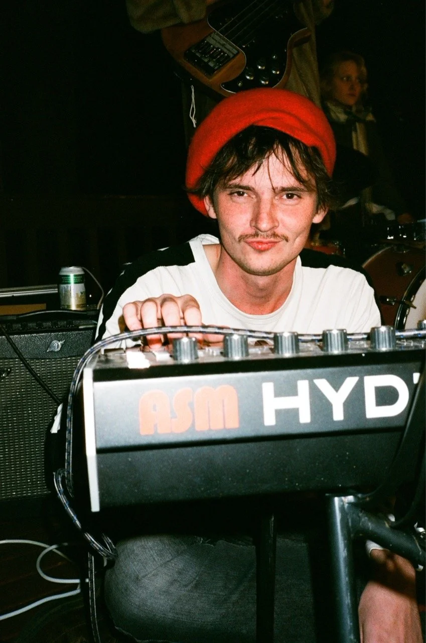 A young man wearing a red hat is sitting behind a music mixing device labeled 'ASM HYD' with a guitar hanging in the background. He is smiling slightly and looking at the camera.