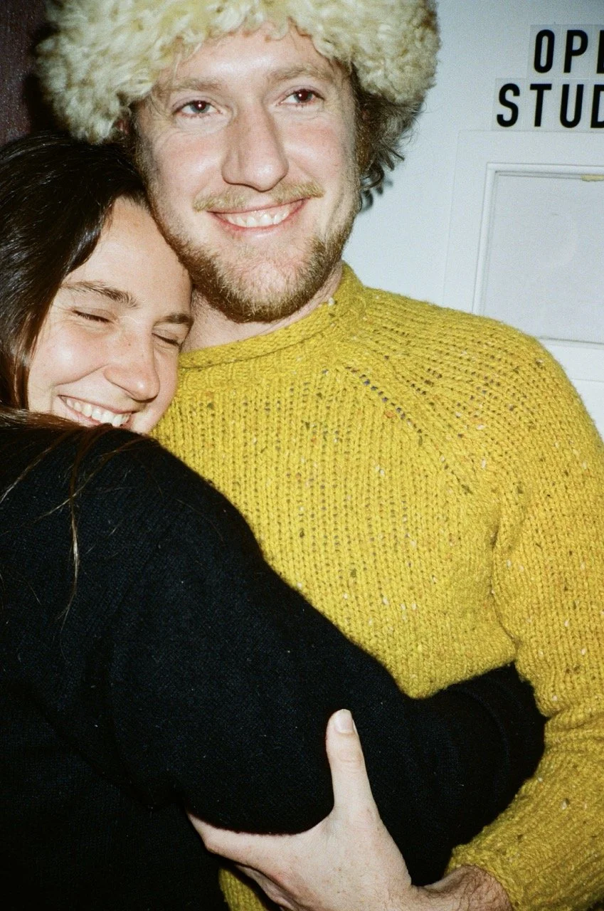 A young man with a beard and a yellow knit sweater, smiling and hugging a young woman with long dark hair and a black sweater, who is also smiling with her eyes closed.